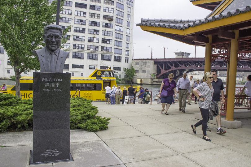 A bronze bust statue at Ping Tom Memorial Park in Chinatown with a yellow Chicago Water Taxi and commuters in the background."