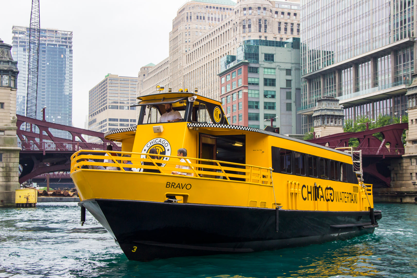 A close-up photo of the yellow and black 'Bravo' Chicago Water Taxi with checkered trim, traveling on a river with a cityscape background including old brick buildings and a bridge.